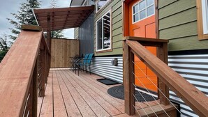 A wooden deck with a metal railing, a covered seating area, and a bright orange door. - Orange Poppy - Seward Alaska Modern Forest Tiny Home Retreat Near Kenai Fjords (Seward)