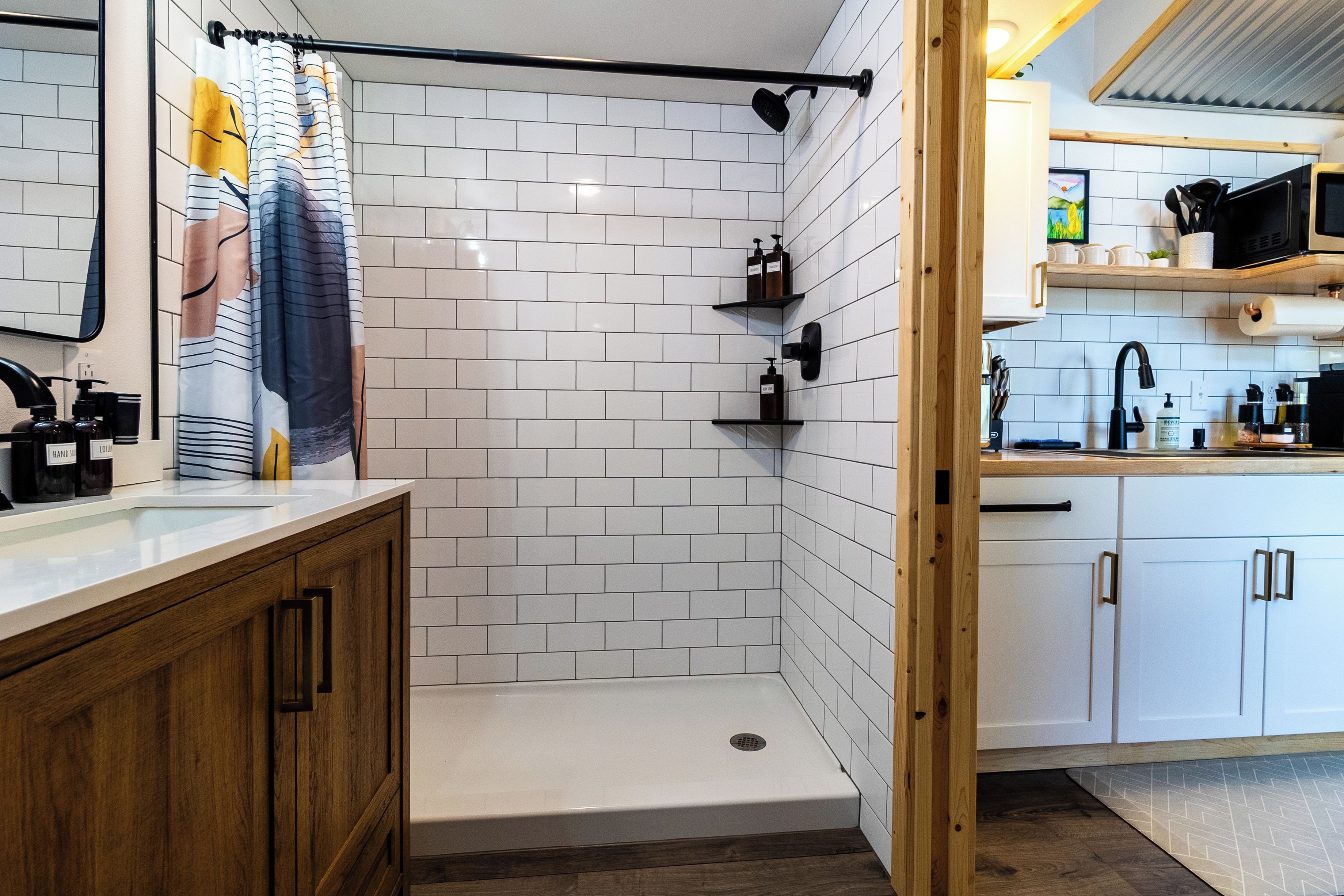 A modern bathroom with white subway tiles, a shower area, wooden cabinets, and a kitchen area with white cabinets and a black faucet.