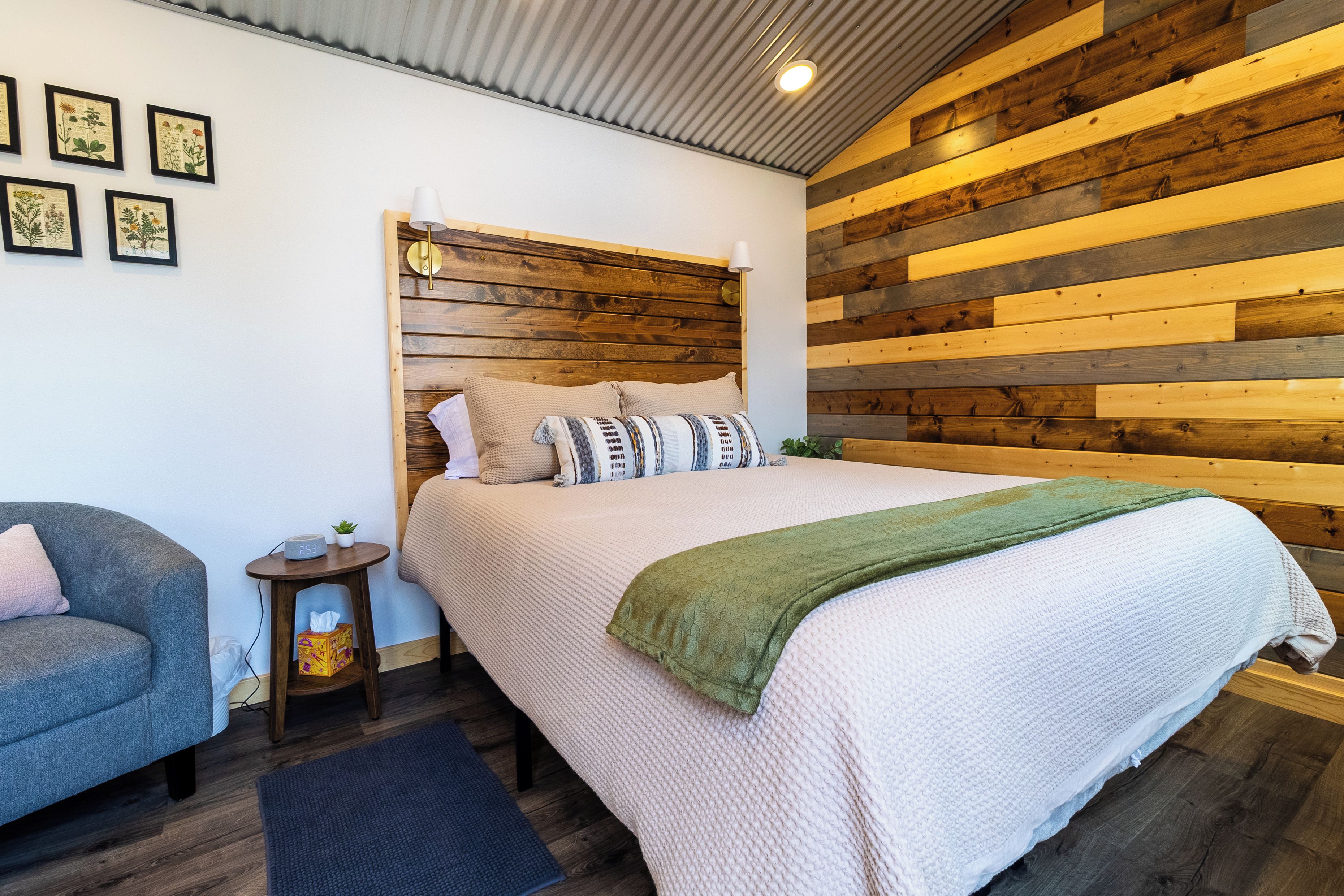 Un dormitorio con cabecera de madera, cama con edredón blanco y almohadas, mesita de noche con lámpara y un sillón gris.
