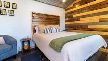 Un dormitorio con cabecera de madera, cama con edredón blanco y almohadas, mesita de noche con lámpara y un sillón gris.