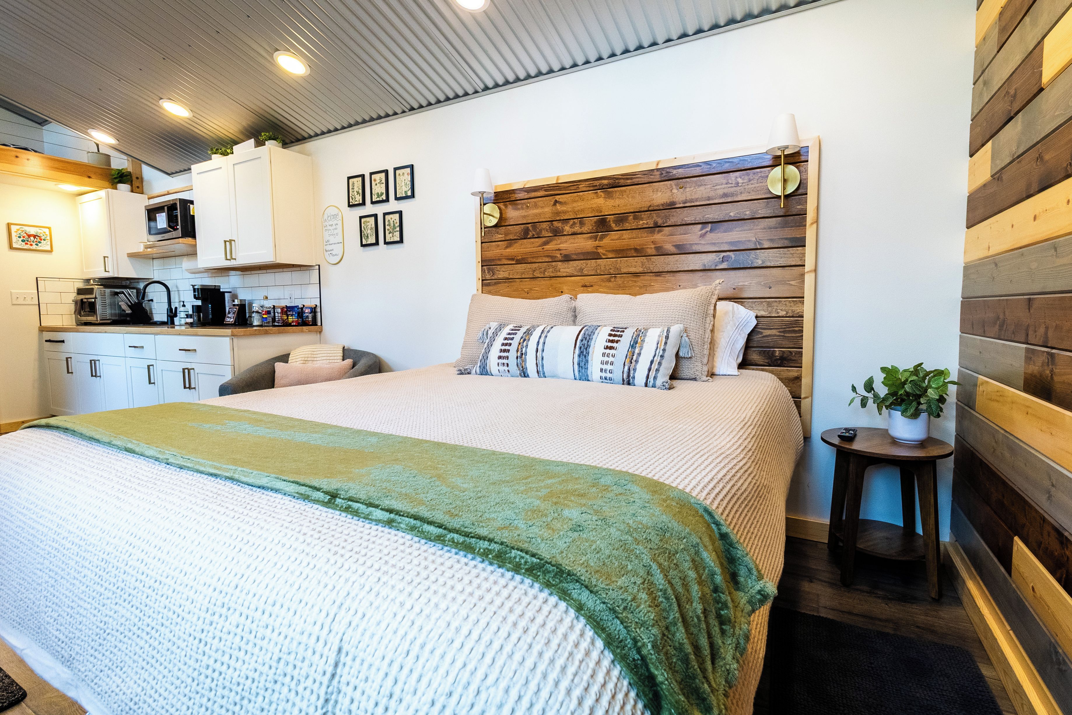 Una camera da letto moderna con una testiera in legno, un letto con molti cuscini, un comodino con una pianta e, sullo sfondo, un angolo cottura.