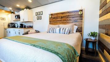 Una camera da letto moderna con una testiera in legno, un letto con molti cuscini, un comodino con una pianta e, sullo sfondo, un angolo cottura.