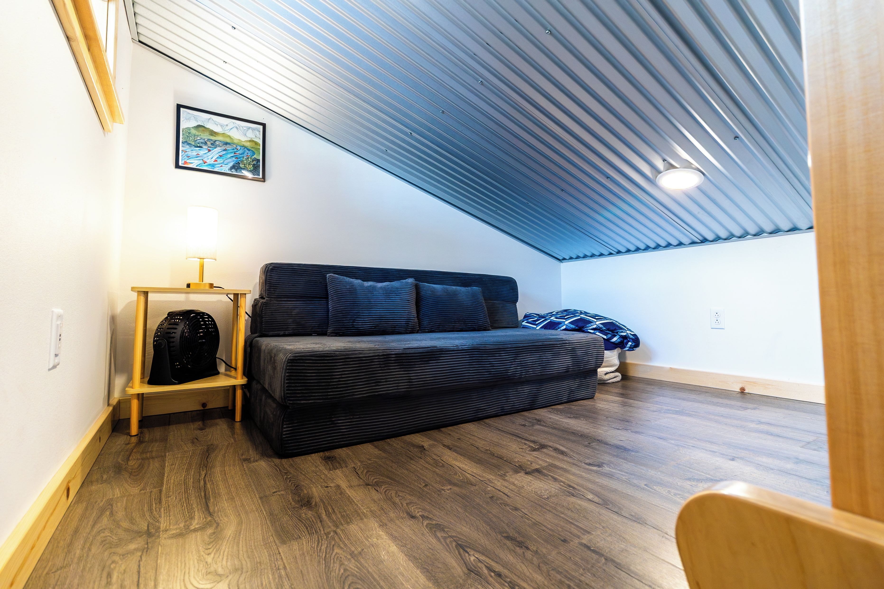 Et lille, hyggeligt rum med skråvægge, en mørk sofa, et lille bord med en lampe og et indrammet billede på væggen.