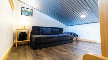 Et lille, hyggeligt rum med skråvægge, en mørk sofa, et lille bord med en lampe og et indrammet billede på væggen.