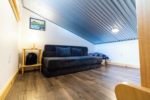 A small, cozy room with a sloped ceiling, a dark sofa, a small table with a lamp, and a framed picture on the wall. - Orange Poppy - Seward Alaska Modern Forest Tiny Home Retreat Near Kenai Fjords (Seward)