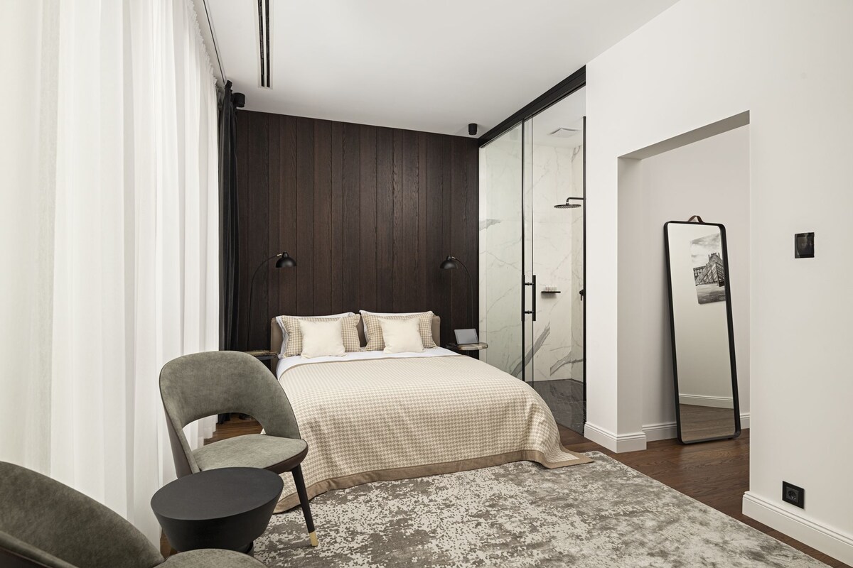 A modern bedroom with a large bed, a chair, a small table, a mirror, and a wardrobe.