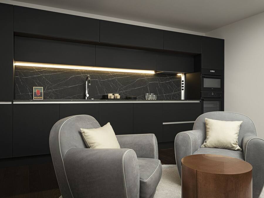 A modern kitchen with dark cabinetry, a marble backsplash, and a built-in oven. There are two grey armchairs with white cushions and a round wooden side table.