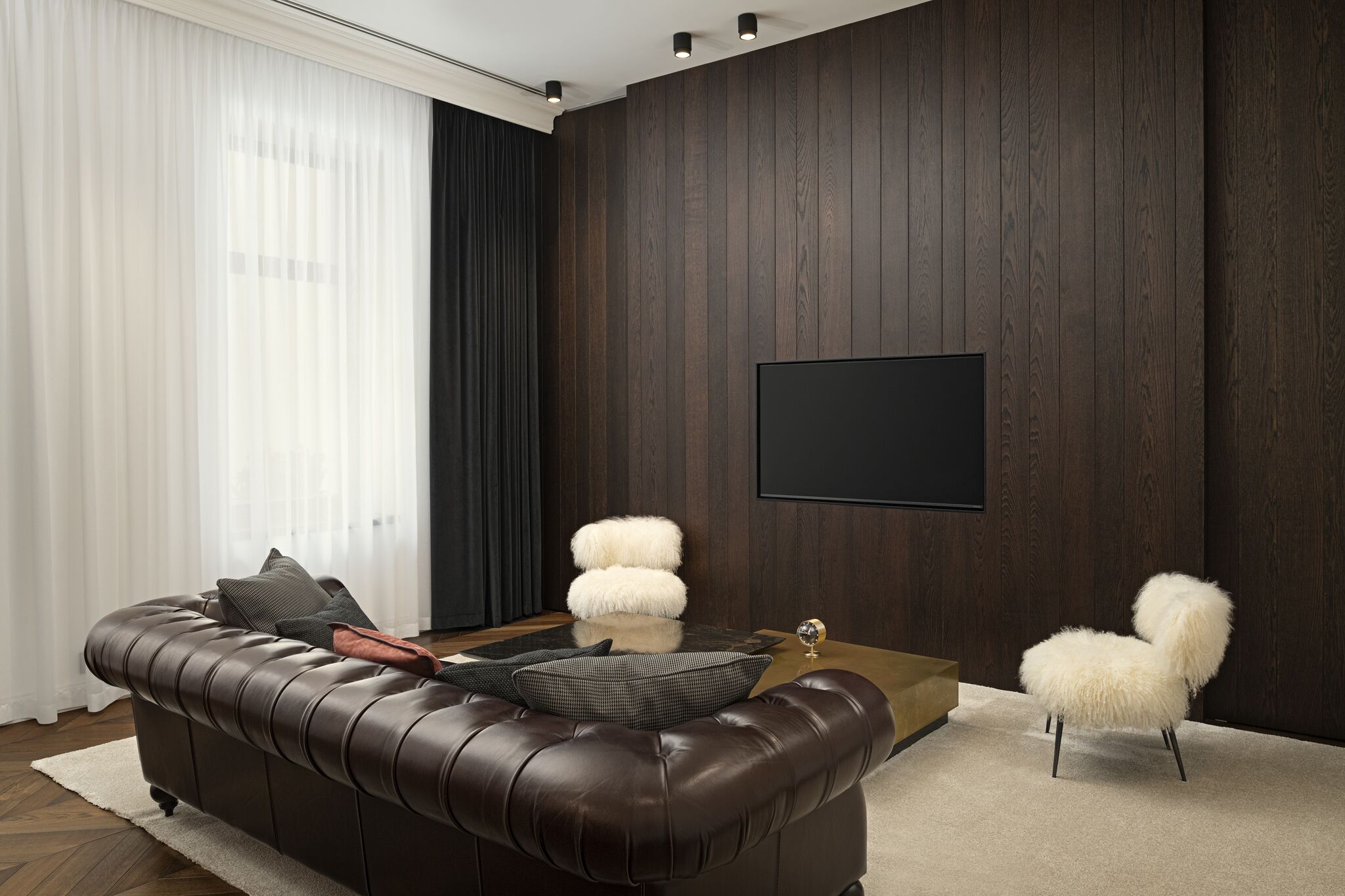 A modern living room with a dark wood-paneled wall, a large flat-screen TV, a leather sofa, and fluffy white chairs.