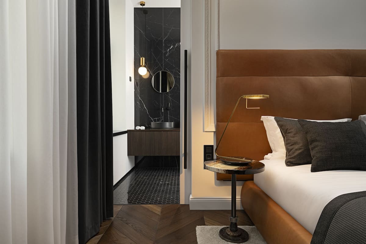 A modern hotel room with a large bed, a bedside table, a lamp, and a view of a bathroom with a black marble wall and a round mirror.