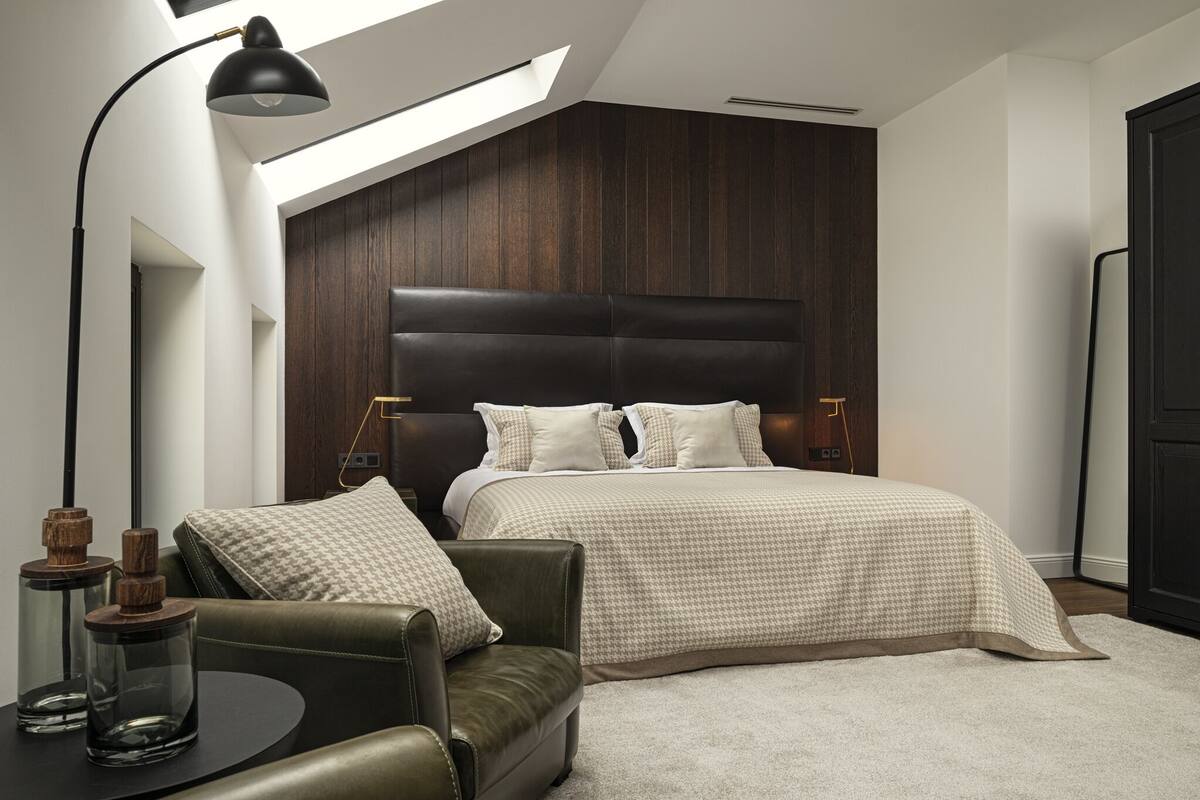 A modern bedroom with a large bed, a skylight, a leather armchair, and a side table with candles.