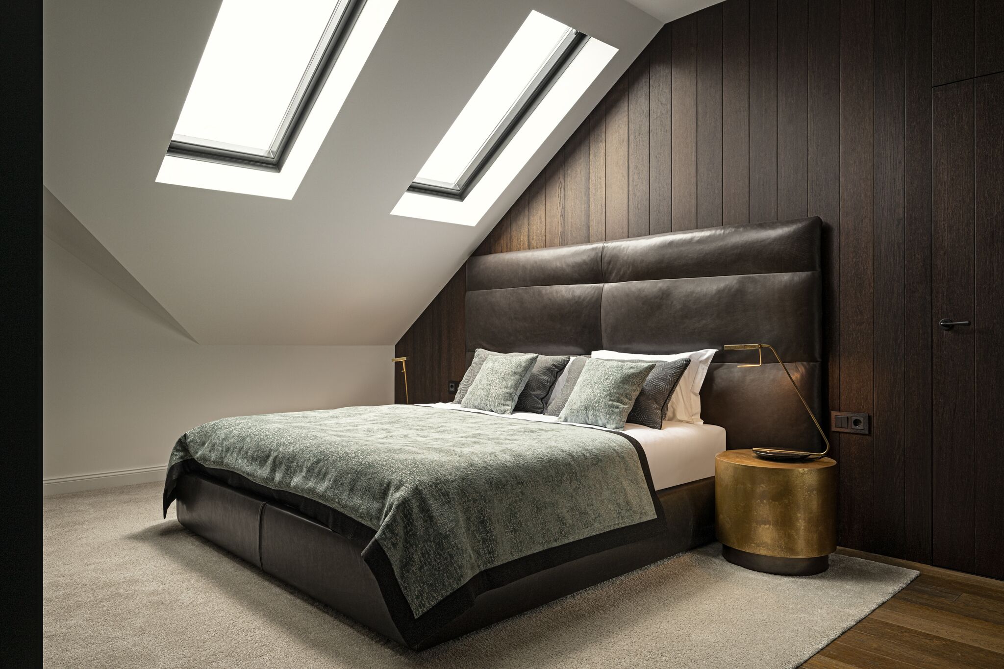 A bedroom with a sloped ceiling, skylights, a large bed, a bedside lamp, and a wooden headboard.