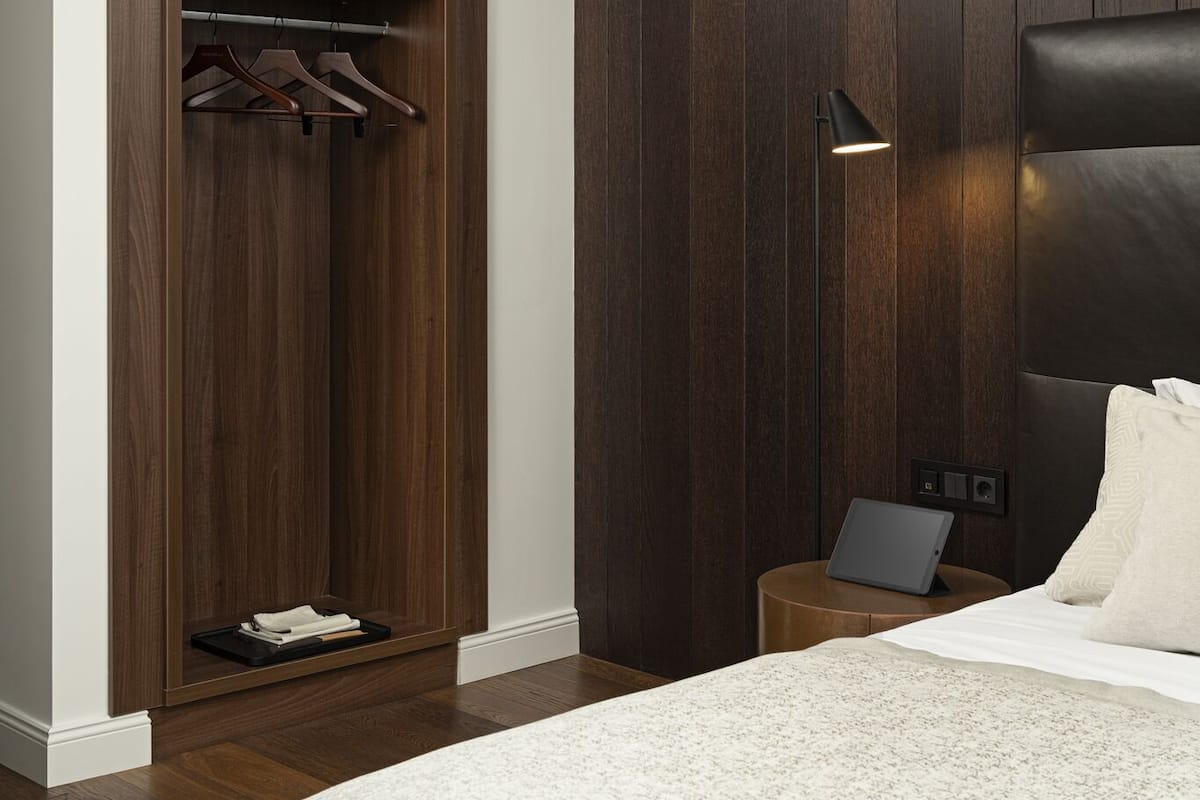 A bedroom with a bed, a wooden wardrobe, a bedside table with a laptop, and a floor lamp.