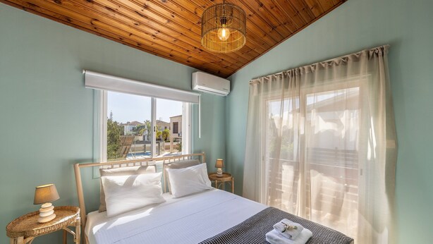 A bedroom with a bed, a window with sheer curtains, a wooden ceiling, and a wall-mounted air conditioner. - Larnaca Oneiro Seaside Villa (Pervolia)