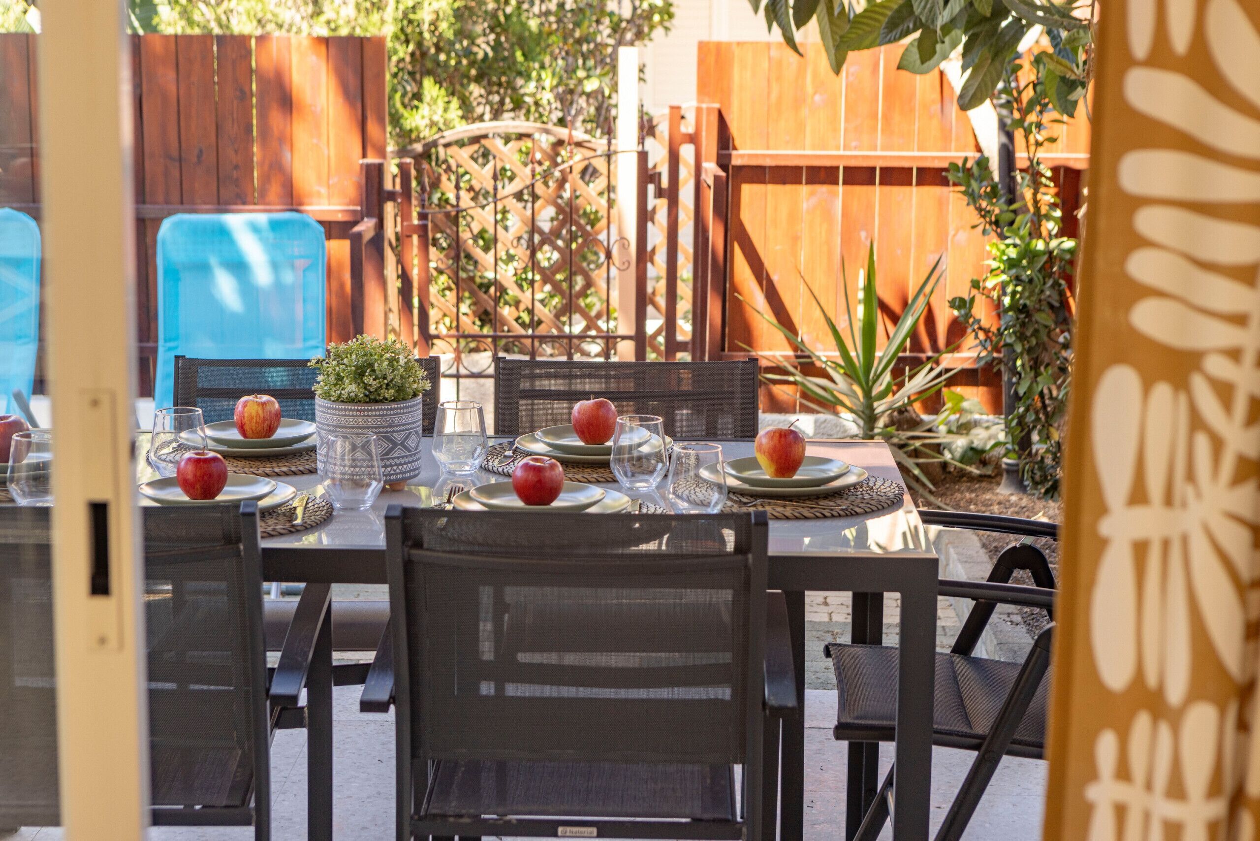 A dining area with a table set for four, surrounded by chairs, with a wooden fence and greenery in the background.