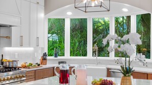 A modern kitchen with white cabinets, a stainless steel stove, and a large window with a tropical view. - Riverfront Riviera Outdoor TV's/Hotub/Tanning Deck (Cape Coral)