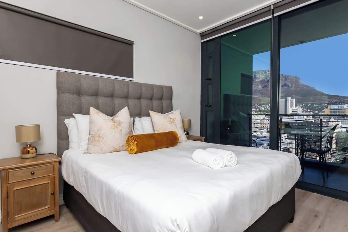 A modern hotel room with a large bed, a nightstand, and a view of a cityscape through a large window.