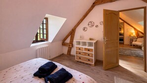 Een slaapkamer met een schuine kap, een bed met een bloemenbedekking, een dressoir met laden, een klein raam en een deur naar een andere kamer.