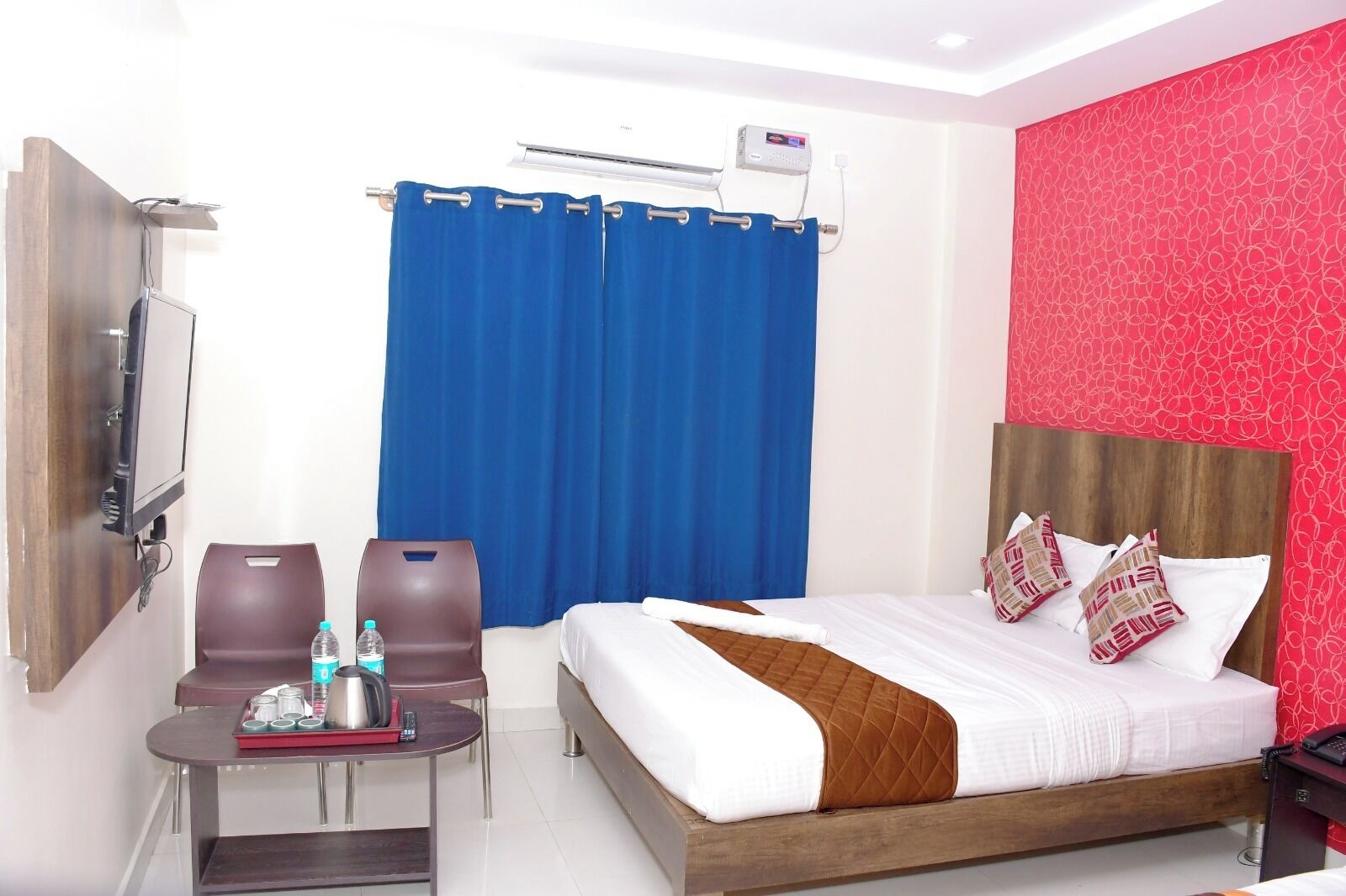 A hotel room with a bed, two chairs, a small table with a bottle and a kettle, a TV, and a red patterned wall.
