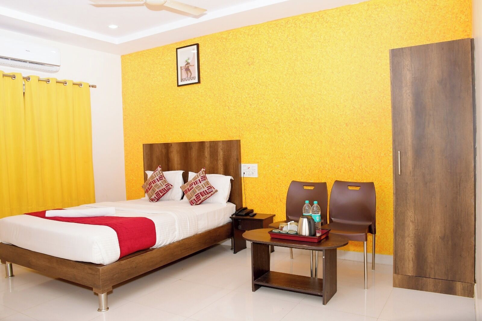 A hotel room with a wooden bed, yellow walls, a ceiling fan, a nightstand, a table with toiletries, and a wardrobe.