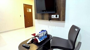 A hotel room with a flat-screen TV mounted on a dark wood panel, a small round table with a tray of bottled water and a remote control, and a black chair. - HARSHA COMFORTS (Chikkamagaluru)