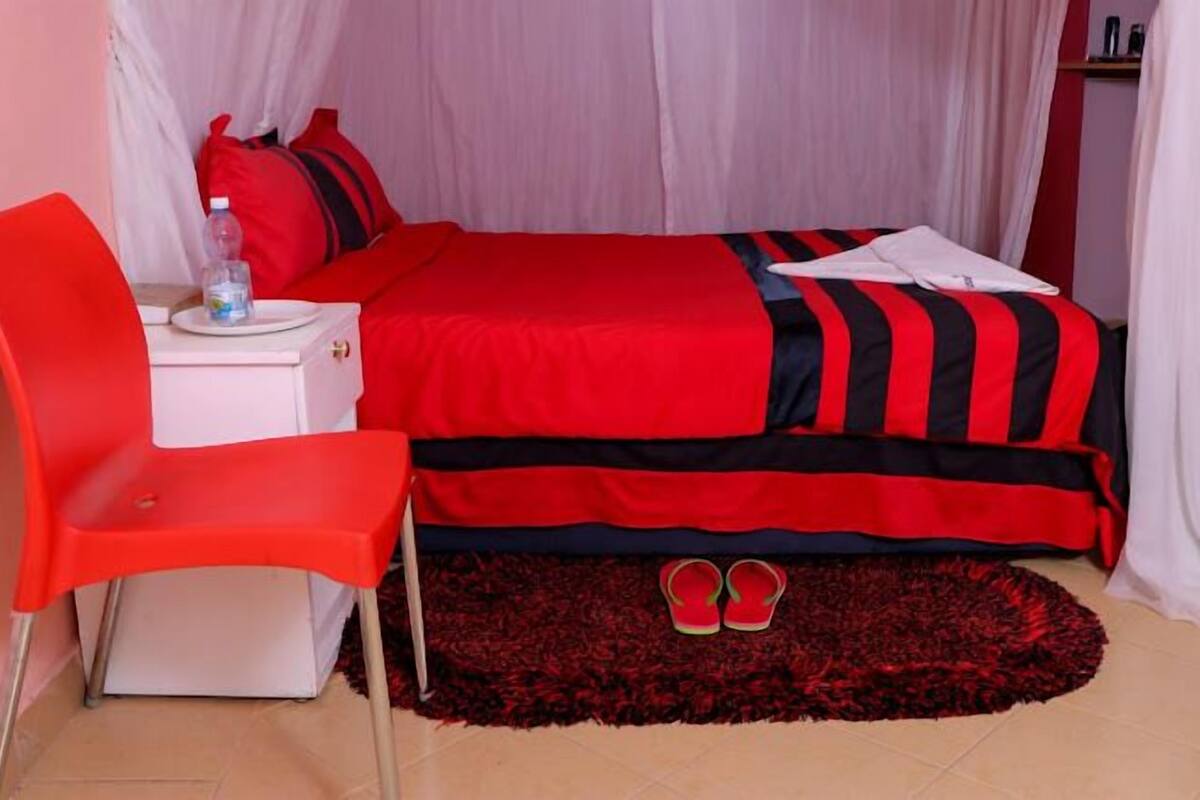 A bedroom with a bed, a red chair, a small table with a water bottle, and a pair of slippers on a rug.
