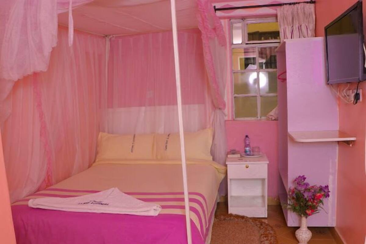 A pink-themed bedroom with a canopy bed, a bedside table with a lamp, and a small white cabinet.