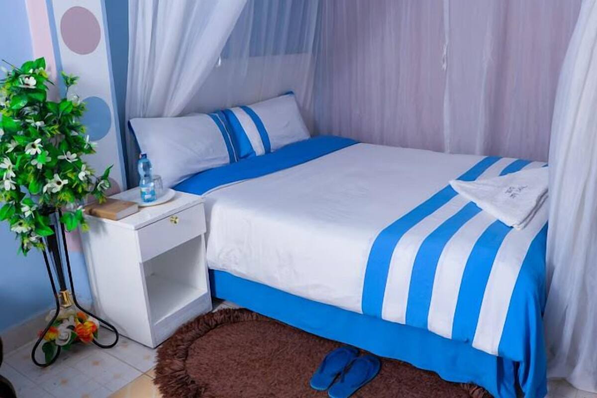 A neatly made bed with blue and white bedding, a bedside table with a water bottle, and a decorative plant.
