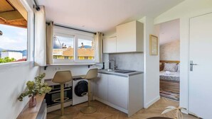 Eine kleine Küche mit Waschmaschine, zwei Barhocker, einem Esstisch und einem Bett im angrenzenden Zimmer.