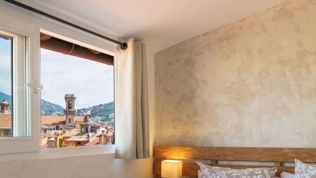 Ein Schlafzimmer mit einem großen Bett, einem Nachttisch mit Lampe, einem Fenster mit Vorhängen und Blick auf Gebäude.