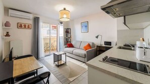 A modern kitchen with a built-in microwave, toaster, and sink. There is a dining area with a table and chairs, and a sofa with cushions. A small side table and a lamp are also visible. - Studio apartment in the Port of Nice (Niza)