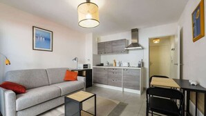 A compact living space with a sofa, coffee table, dining table, chairs, kitchenette, and a hanging light fixture. - Studio apartment in the Port of Nice (Niza)
