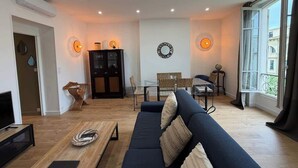 A modern living room with a sofa, dining table, and chairs, a TV, and a large window. - Charming 2-bedroom apartment on Avenue Jean Médecin (Niza)