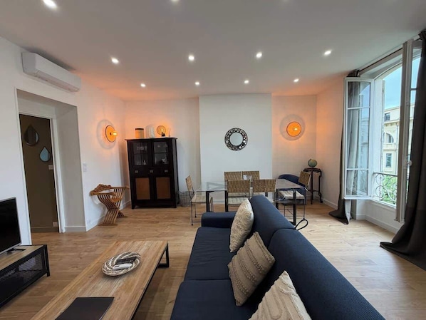 A modern living room with a sofa, dining table, and chairs, a TV, and a large window.