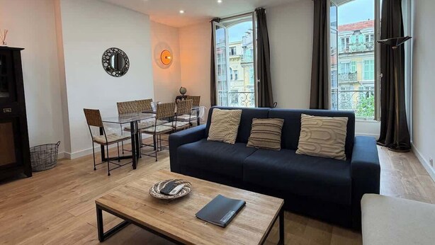 A modern living room with a dark blue sofa, a wooden coffee table, and a dining area with chairs and a table. - Charming 2-bedroom apartment on Avenue Jean Médecin (Niza)
