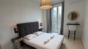 A bedroom with a bed, bedside tables, a desk, a chair, a lamp, a mirror, and a window with curtains. - Charming 2-bedroom apartment on Avenue Jean Médecin (Niza)