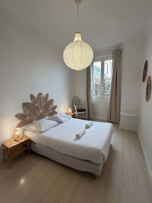 A bedroom with a large bed, a hanging light fixture, a chair, and a window with curtains.