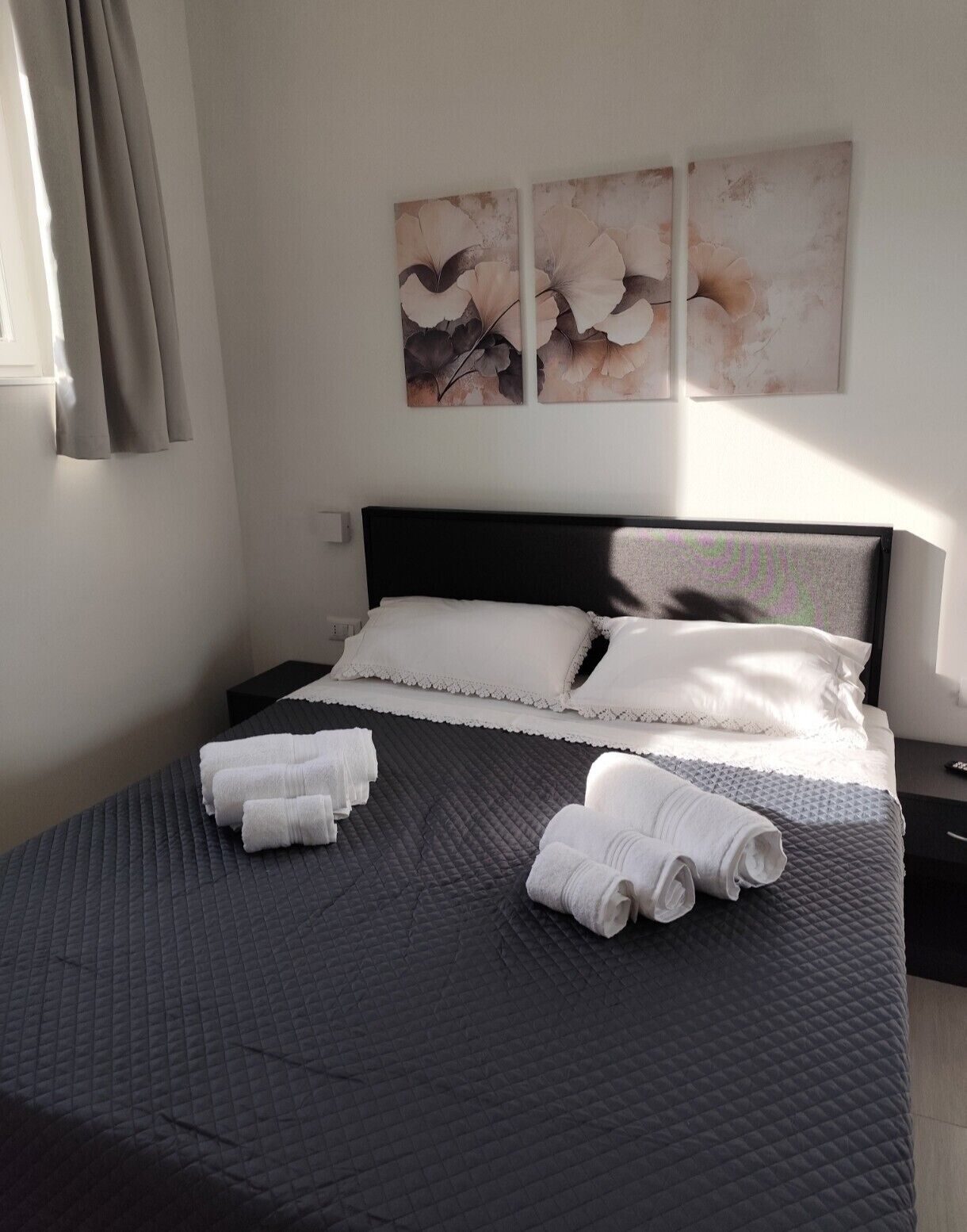 Una cama bien hecha con un edredón oscuro, almohadas blancas y toallas enrolladas sobre un cabecero oscuro.