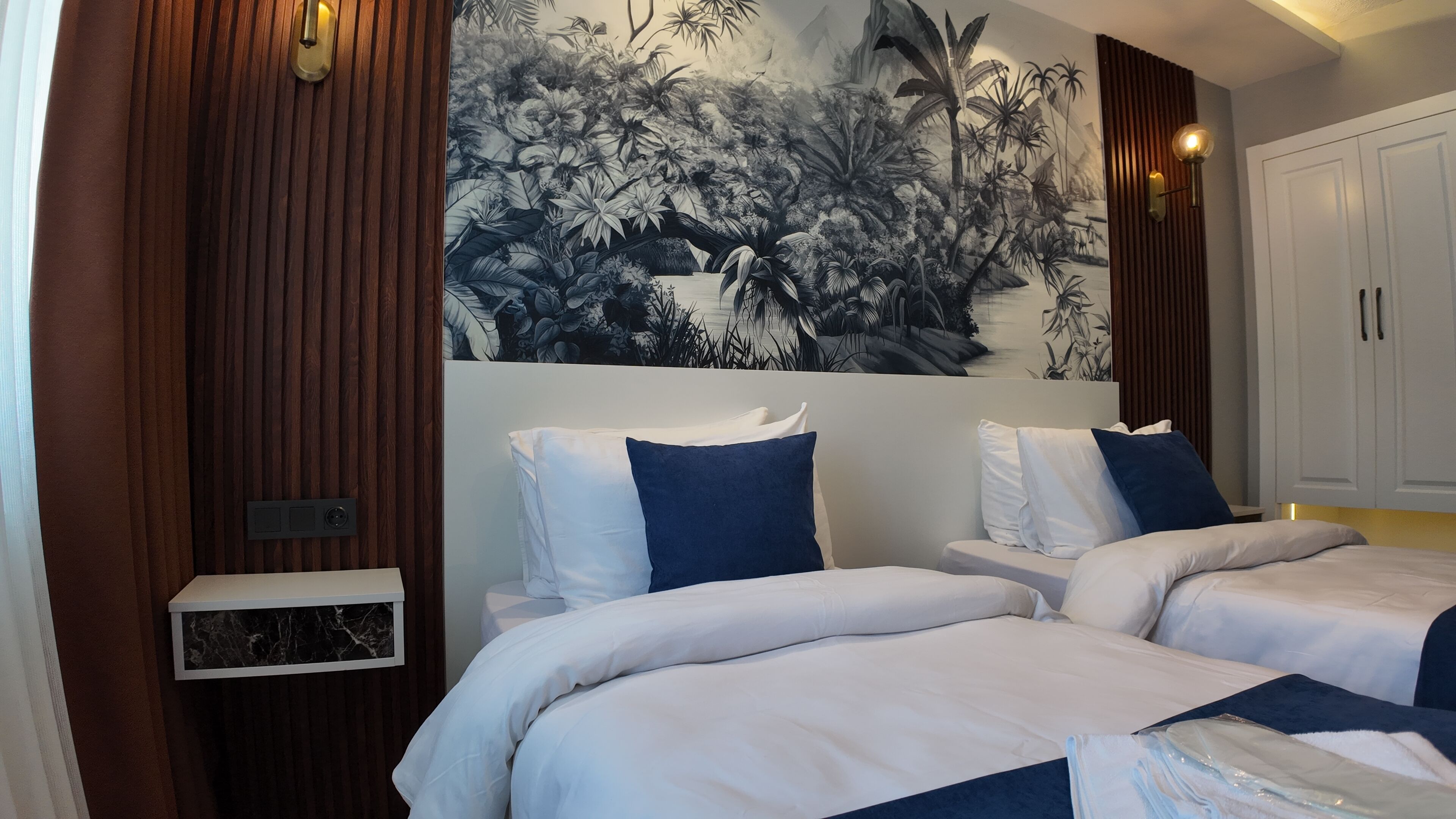 Ein Hotelzimmer mit zwei Betten, einem großen tropischen Wandgemälde, Holzvertäfelung und einem weißen Kleiderschrank.
