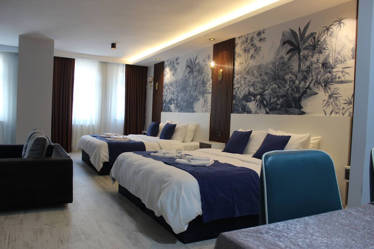 Quarto de hotel com duas camas, um sofá, uma escrivaninha e um grande mural na parede.
