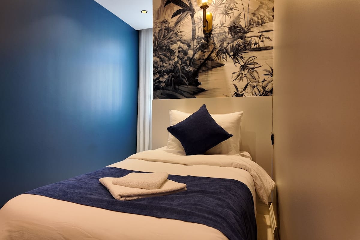 Quarto com cama, mesa de cabeceira, luminária na parede e um mural.