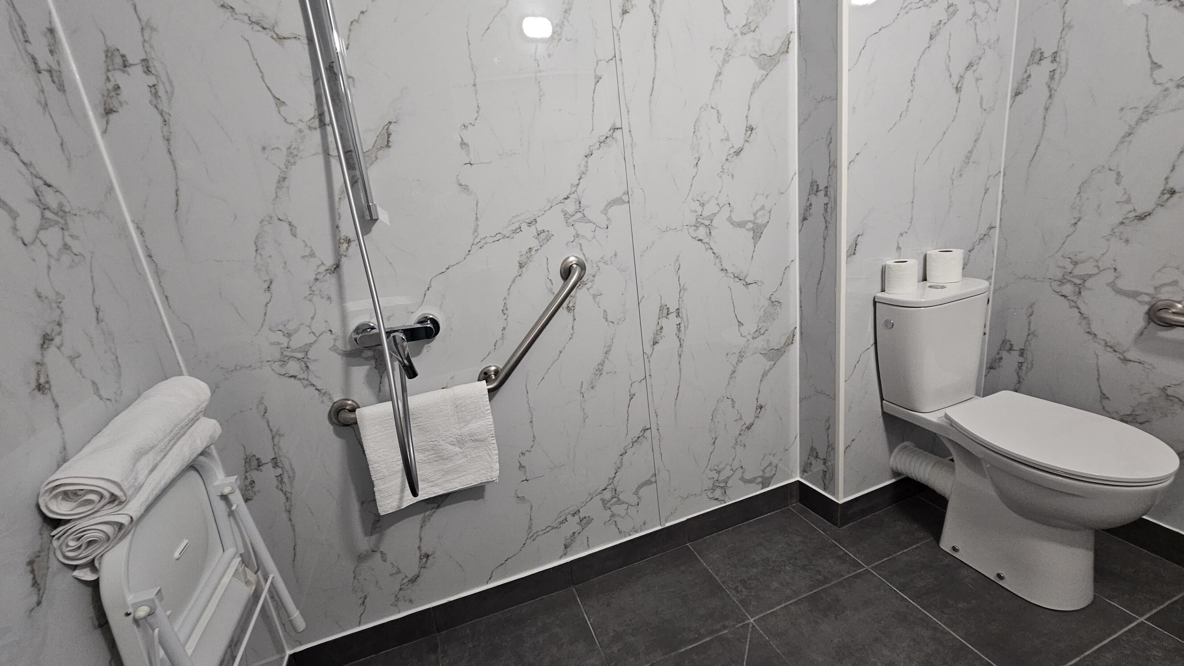 Un baño con pared de mármol, inodoro blanco, barra para toallas y pasamanos.
