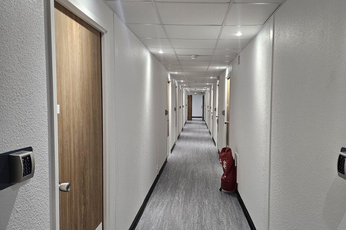 Un pasillo largo y estrecho de hotel con puertas a ambos lados, un extintor a la derecha e iluminación empotrada en el techo.