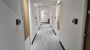 Corredor de hotel com portas em ambos os lados, carpete cinzento e uma janela triangular no final.