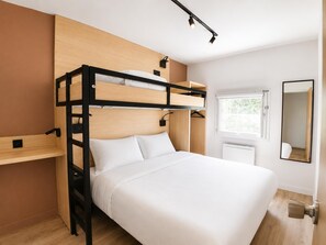 Uma cama beliche com estrutura de madeira, colchão branco e mesa de cabeceira. Há uma janela com vista para o verde lá fora, e um espelho na parede.