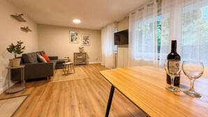 Una sala moderna con piso de madera, un sofá gris, una mesita auxiliar con una planta y una mesa de comedor con copas de vino.