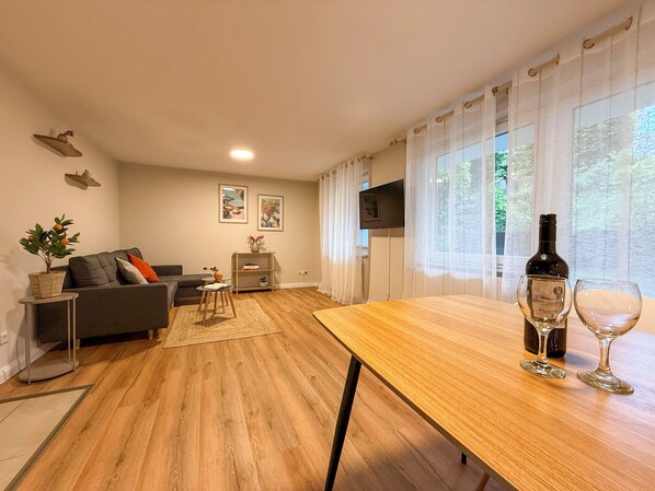 A modern living room with a wooden floor, a grey sofa, a small side table with a plant, and a dining table with wine glasses.