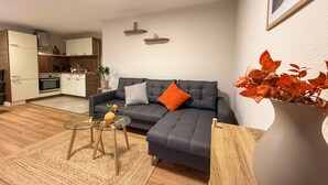 Una sala de estar moderna con un sofá gris, una pequeña mesa de centro de vidrio y una cocina al fondo.