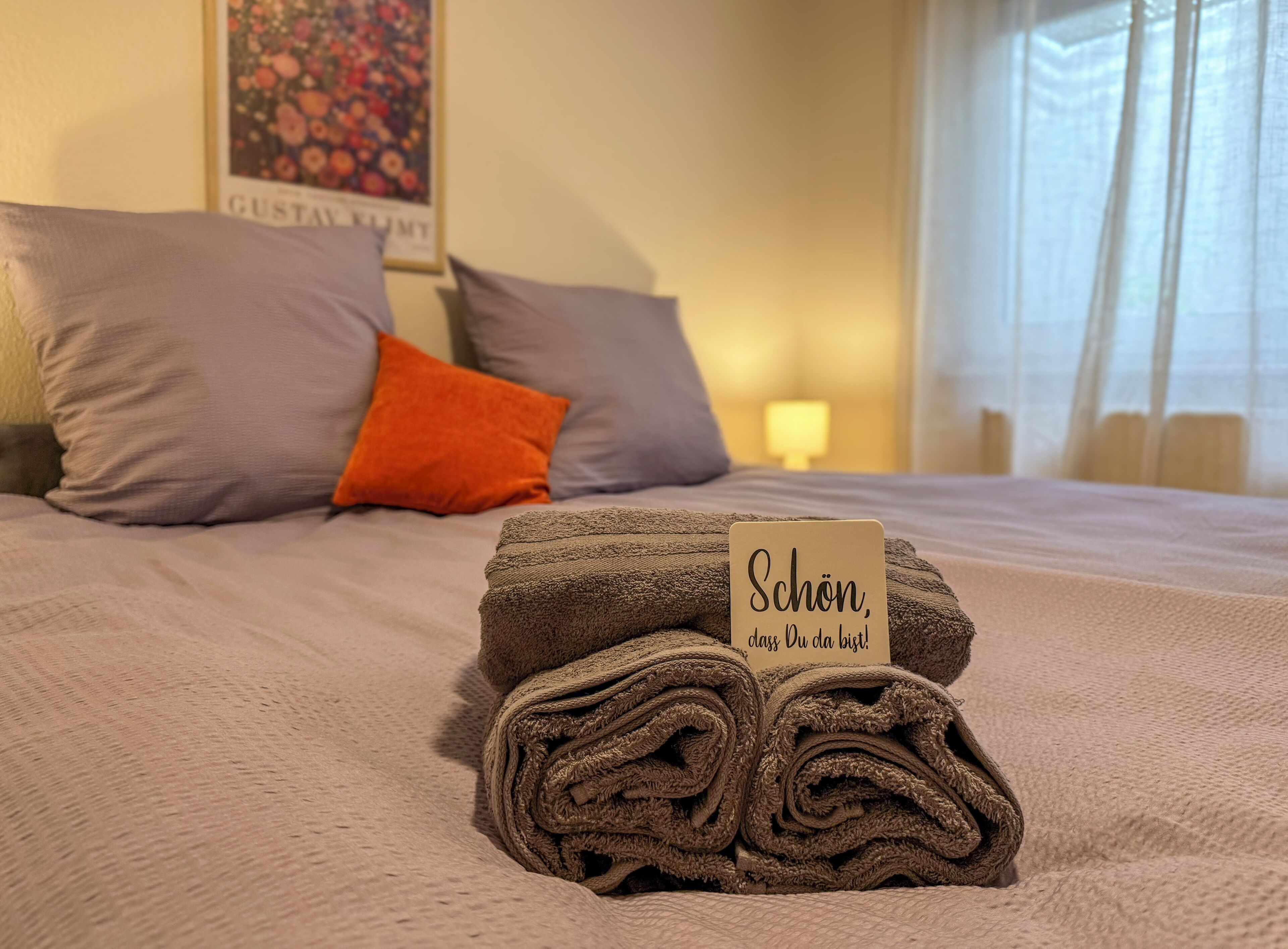 Un lit bien fait, avec des serviettes pliées en rouleaux et une pancarte sur laquelle on peut lire : « Schön, dass du da bist ! » dans une chambre d’hôtel.