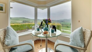 Une pièce d’angle avec une table en verre, deux fauteuils en osier, un vase de fleurs et une vue panoramique sur des champs verdoyants et des collines.
