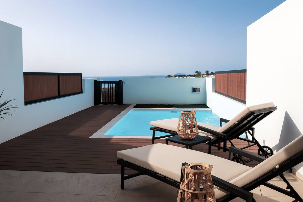 Ein Poolbereich auf dem Dach mit zwei Liegestühlen und einem kleinen Tisch, umgeben von einer Holzterrasse und weißen Wänden.
